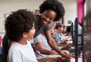 Teacher Helping Male Pupil Line Of High School Students Working at Screens In Computer Class